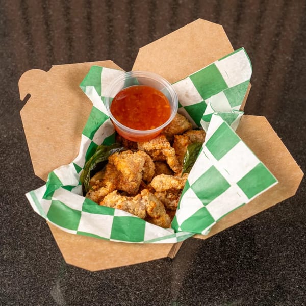 Crispy fried chicken pieces with green leaves and a side of red dipping sauce in a takeout box.