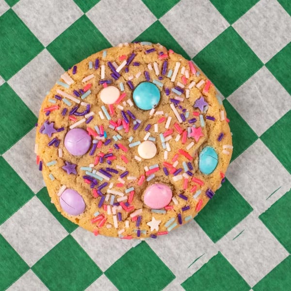 A colorful sugar cookie with sprinkles and candy pieces on green and white checkered paper.