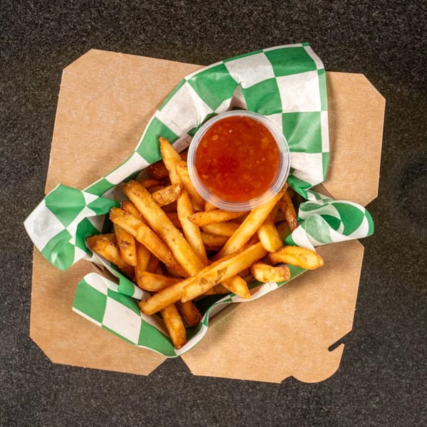 Golden french fries in a brown paper container with green and white checkered lining, served with a side of red dipping sauce.